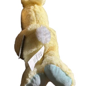 May include: A plush yellow bunny toy with a white fluffy tail and light blue feet. The bunny has a tag attached. The toy is made of soft, fuzzy material.