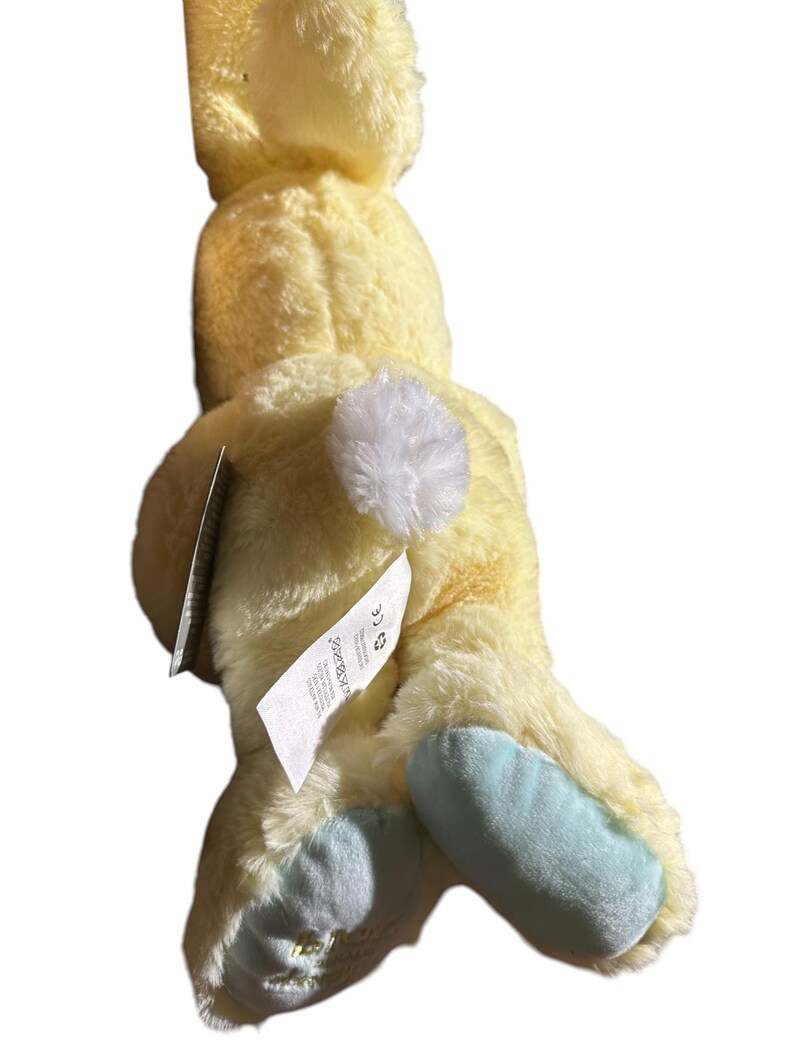 May include: A plush yellow bunny toy with a white fluffy tail and light blue feet. The bunny has a tag attached. The toy is made of soft, fuzzy material.