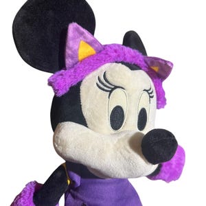 K&ouml;nnte beinhalten: Ein Pl&uuml;sch Minnie Mouse-Puppe in einem Halloween-Kost&uuml;m. Die Puppe hat einen schwarzen Kopf und Ohren, ein wei&szlig;es Gesicht und ein lila Outfit mit einem orangefarbenen Aufn&auml;her. Die Puppe tr&auml;gt ein lila Stirnband mit Katzenohren.