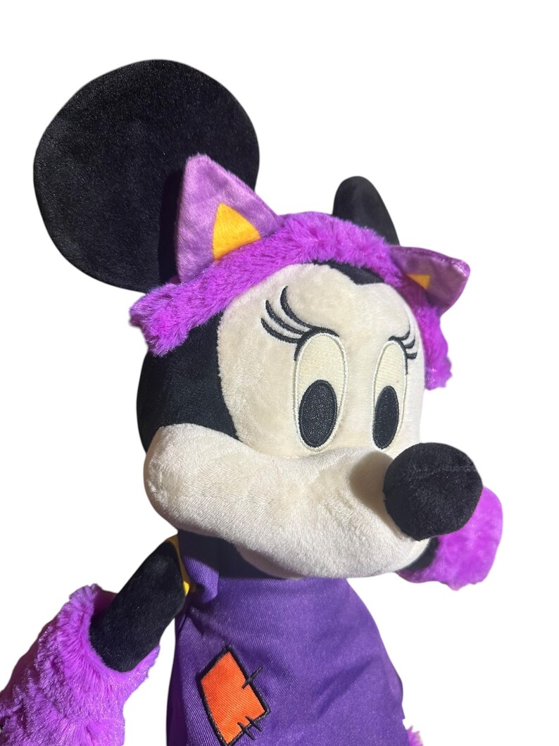 K&ouml;nnte beinhalten: Ein Pl&uuml;sch Minnie Mouse-Puppe in einem Halloween-Kost&uuml;m. Die Puppe hat einen schwarzen Kopf und Ohren, ein wei&szlig;es Gesicht und ein lila Outfit mit einem orangefarbenen Aufn&auml;her. Die Puppe tr&auml;gt ein lila Stirnband mit Katzenohren.