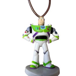 May include: A Buzz Lightyear pendant necklace. The pendant is a miniature figurine of the character from the Toy Story movie franchise. The figure is white, green, and purple, with a brown cord necklace.