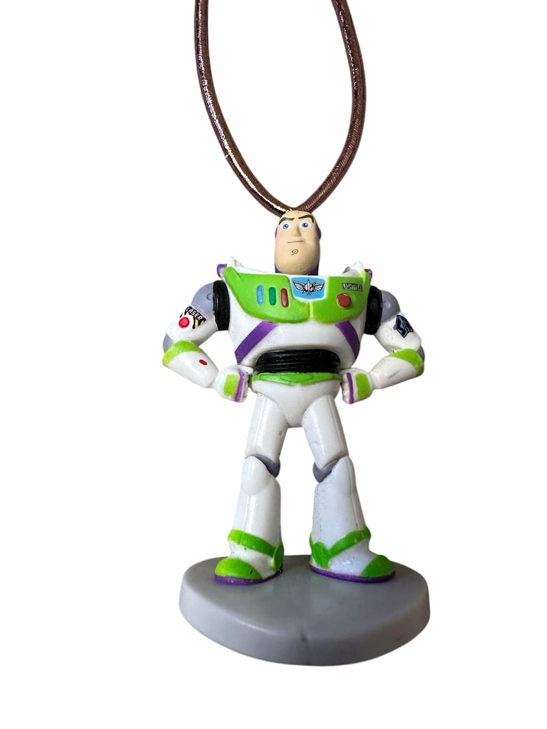 May include: A Buzz Lightyear pendant necklace. The pendant is a miniature figurine of the character from the Toy Story movie franchise. The figure is white, green, and purple, with a brown cord necklace.