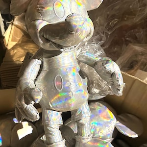Puede incluir: Un peluche de Mickey Mouse plateado con un acabado hologr&aacute;fico iridiscente. El peluche tiene orejas grandes, una cara redonda y las caracter&iacute;sticas cl&aacute;sicas de Mickey Mouse. El juguete est&aacute; en una caja con otros peluches similares.
