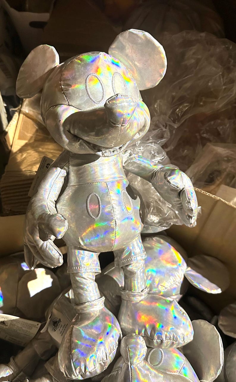 Puede incluir: Un peluche de Mickey Mouse plateado con un acabado hologr&aacute;fico iridiscente. El peluche tiene orejas grandes, una cara redonda y las caracter&iacute;sticas cl&aacute;sicas de Mickey Mouse. El juguete est&aacute; en una caja con otros peluches similares.