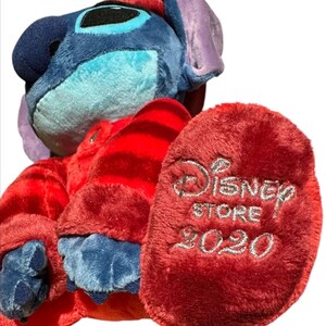 May include: A blue plush character with large ears wearing a red long-sleeved shirt with horizontal stripes. The plush has dark blue claws and a red foot pad embroidered with the silver text Disney STORE 2020. The character has a light blue face.