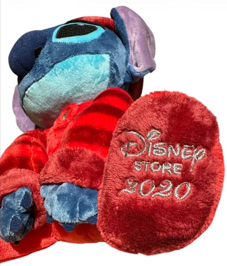 May include: A blue plush character with large ears wearing a red long-sleeved shirt with horizontal stripes. The plush has dark blue claws and a red foot pad embroidered with the silver text Disney STORE 2020. The character has a light blue face.