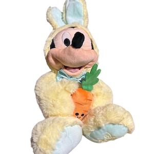 May include: A plush Mickey Mouse dressed in a yellow bunny costume with light blue accents. Mickey holds a smiling orange carrot with green leaves. The plush toy has a light blue bow tie and is set against a white background.