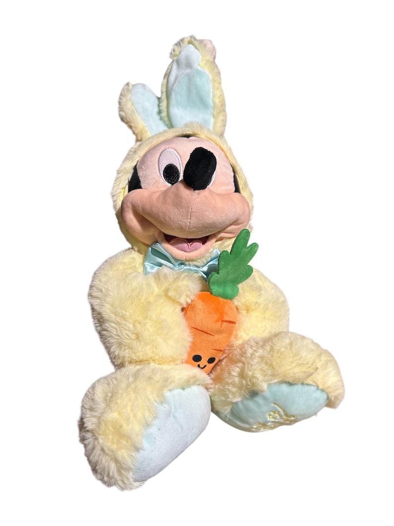 May include: A plush Mickey Mouse dressed in a yellow bunny costume with light blue accents. Mickey holds a smiling orange carrot with green leaves. The plush toy has a light blue bow tie and is set against a white background.