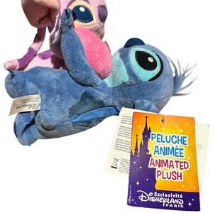May include: Two plush toys featuring the characters Stitch and Angel. The blue Stitch plush is in the foreground, while the pink Angel plush is positioned behind it. A rectangular tag attached to the blue plush reads PELUCHE ANIM&Eacute;E ANIMATED PLUSH.
