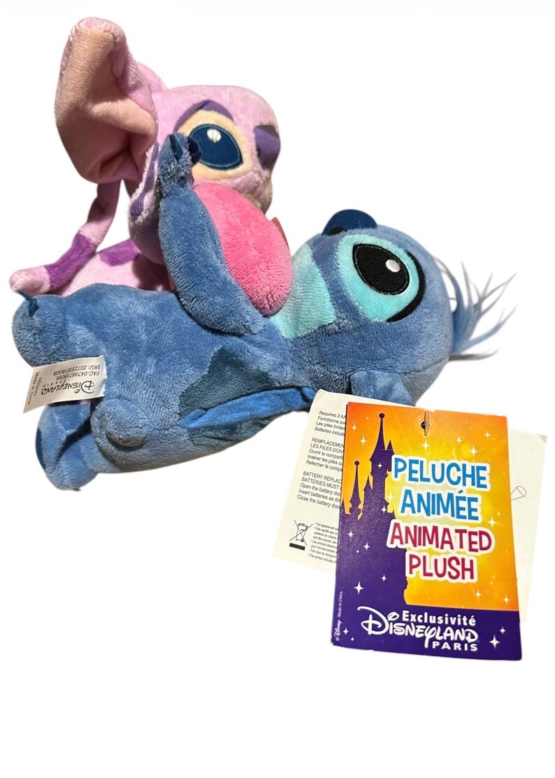 May include: Two plush toys featuring the characters Stitch and Angel. The blue Stitch plush is in the foreground, while the pink Angel plush is positioned behind it. A rectangular tag attached to the blue plush reads PELUCHE ANIM&Eacute;E ANIMATED PLUSH.