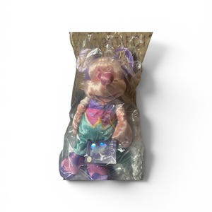 May include: A plush toy mouse in a clear plastic bag. The mouse has pink fur, purple ears, and a colorful outfit with a heart design. A tag with the words "Mickey Mouse" is attached to the toy.