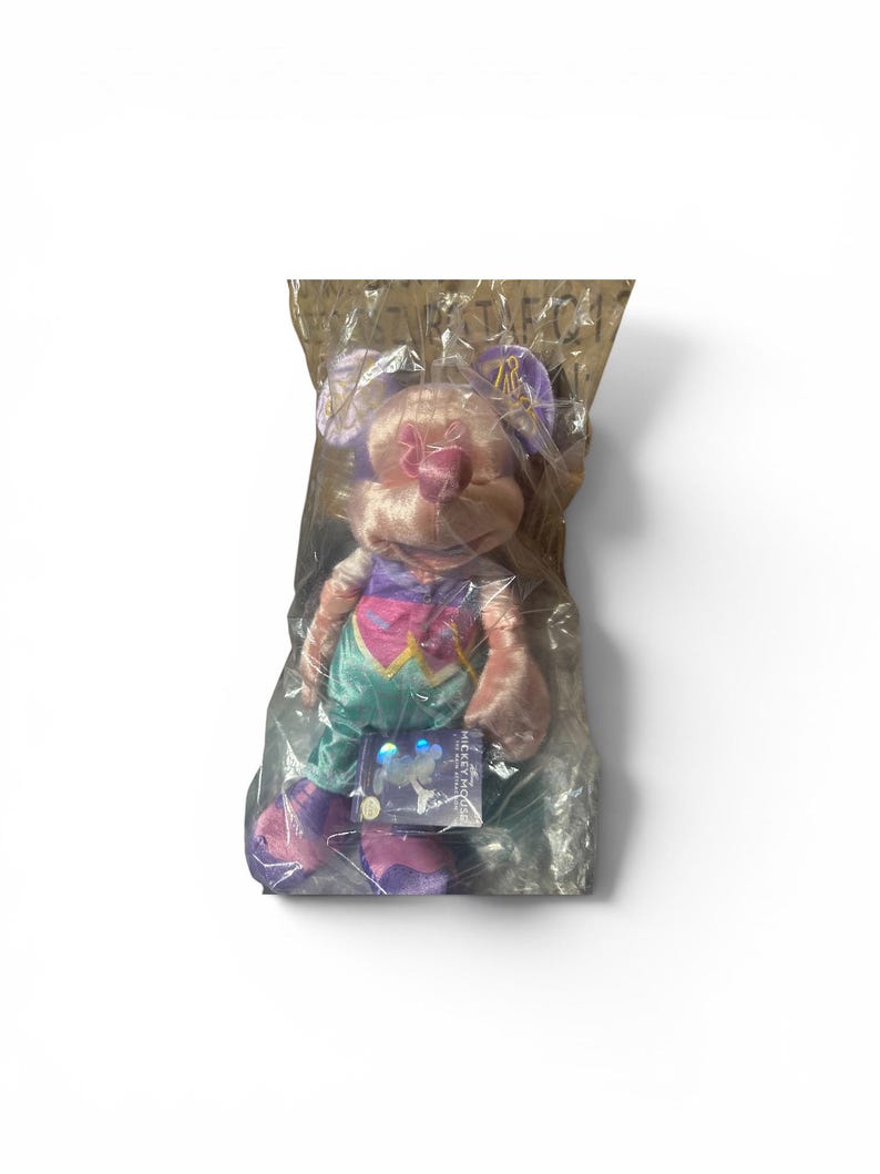May include: A plush toy mouse in a clear plastic bag. The mouse has pink fur, purple ears, and a colorful outfit with a heart design. A tag with the words "Mickey Mouse" is attached to the toy.