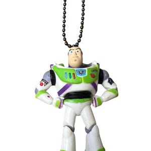 Puede incluir: Una figura de Buzz Lightyear, con un dise&ntilde;o de traje espacial blanco y verde con detalles morados. El juguete tiene una cadena negra para colgar y se apoya sobre una base gris. La figura tiene una expresi&oacute;n decidida.