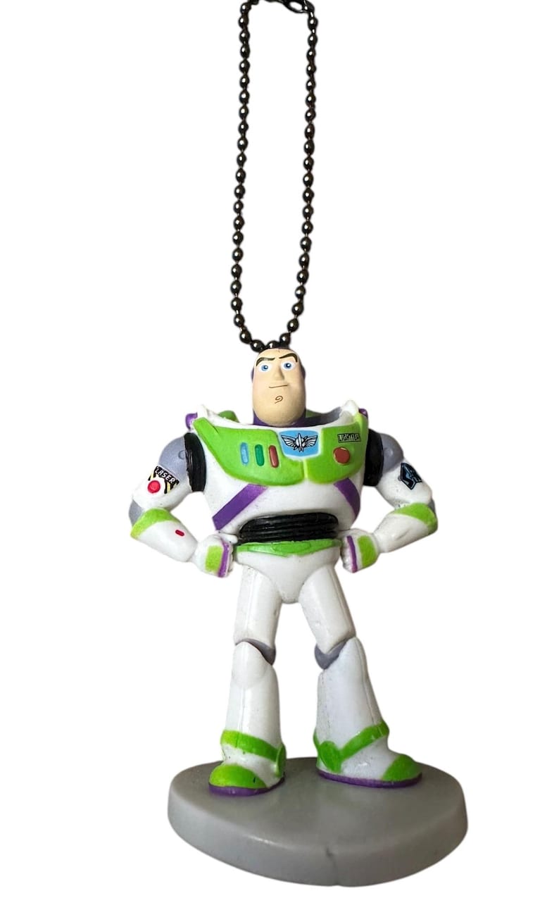 Puede incluir: Una figura de Buzz Lightyear, con un dise&ntilde;o de traje espacial blanco y verde con detalles morados. El juguete tiene una cadena negra para colgar y se apoya sobre una base gris. La figura tiene una expresi&oacute;n decidida.