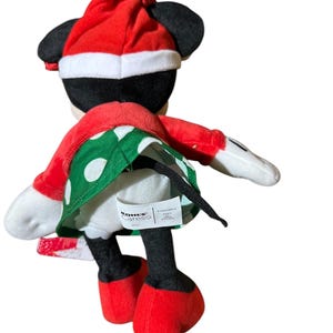 May include: A plush Minnie Mouse doll wearing a red Santa hat and a red and green polka dot dress. The doll has black ears, black legs, and red shoes. The doll is facing away from the camera.