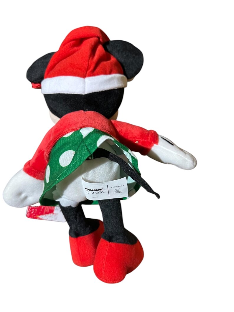 May include: A plush Minnie Mouse doll wearing a red Santa hat and a red and green polka dot dress. The doll has black ears, black legs, and red shoes. The doll is facing away from the camera.