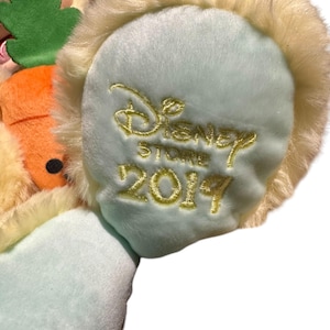 May include: Close-up of a plush toy's ear, featuring a light blue base with the embroidered words "Disney Store 2019" in gold. The ear is trimmed with soft, cream-colored fur, suggesting a character from a Disney franchise.