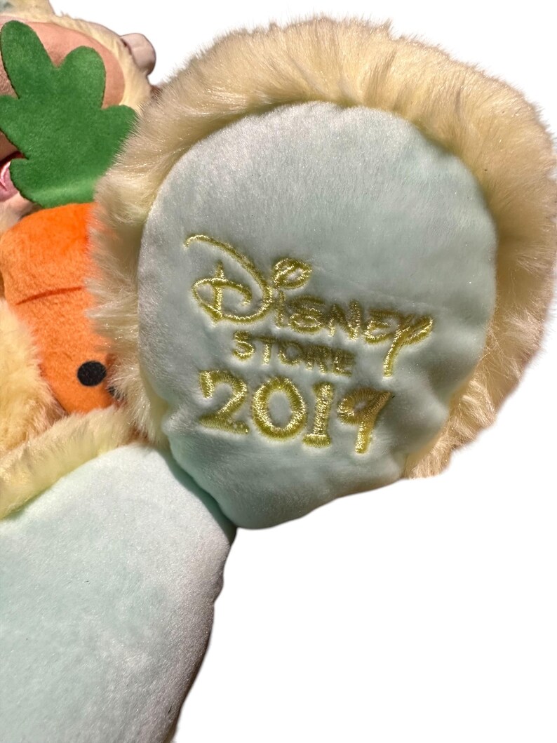 May include: Close-up of a plush toy's ear, featuring a light blue base with the embroidered words "Disney Store 2019" in gold. The ear is trimmed with soft, cream-colored fur, suggesting a character from a Disney franchise.