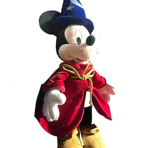 May include: A plush Mickey Mouse dressed as the Sorcerer's Apprentice from Fantasia. Mickey wears a blue wizard hat with silver stars and a red velvet robe with a gold rope tie. His gloves are white, and his shoes are tan.