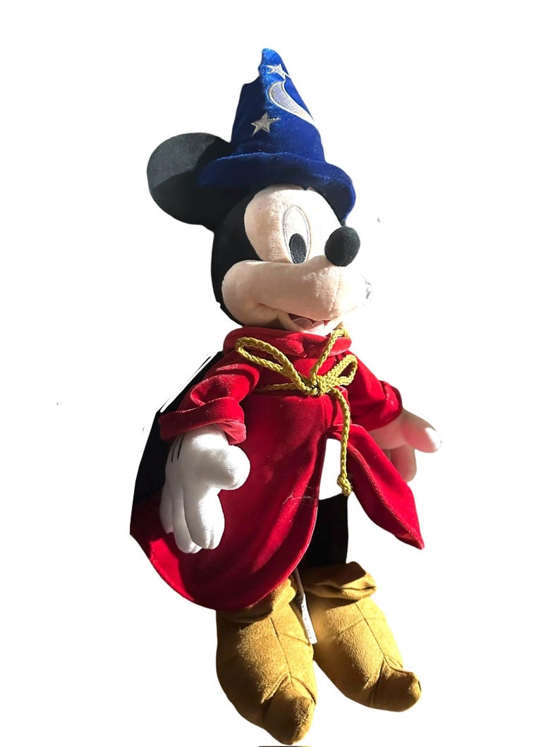 May include: A plush Mickey Mouse dressed as the Sorcerer's Apprentice from Fantasia. Mickey wears a blue wizard hat with silver stars and a red velvet robe with a gold rope tie. His gloves are white, and his shoes are tan.