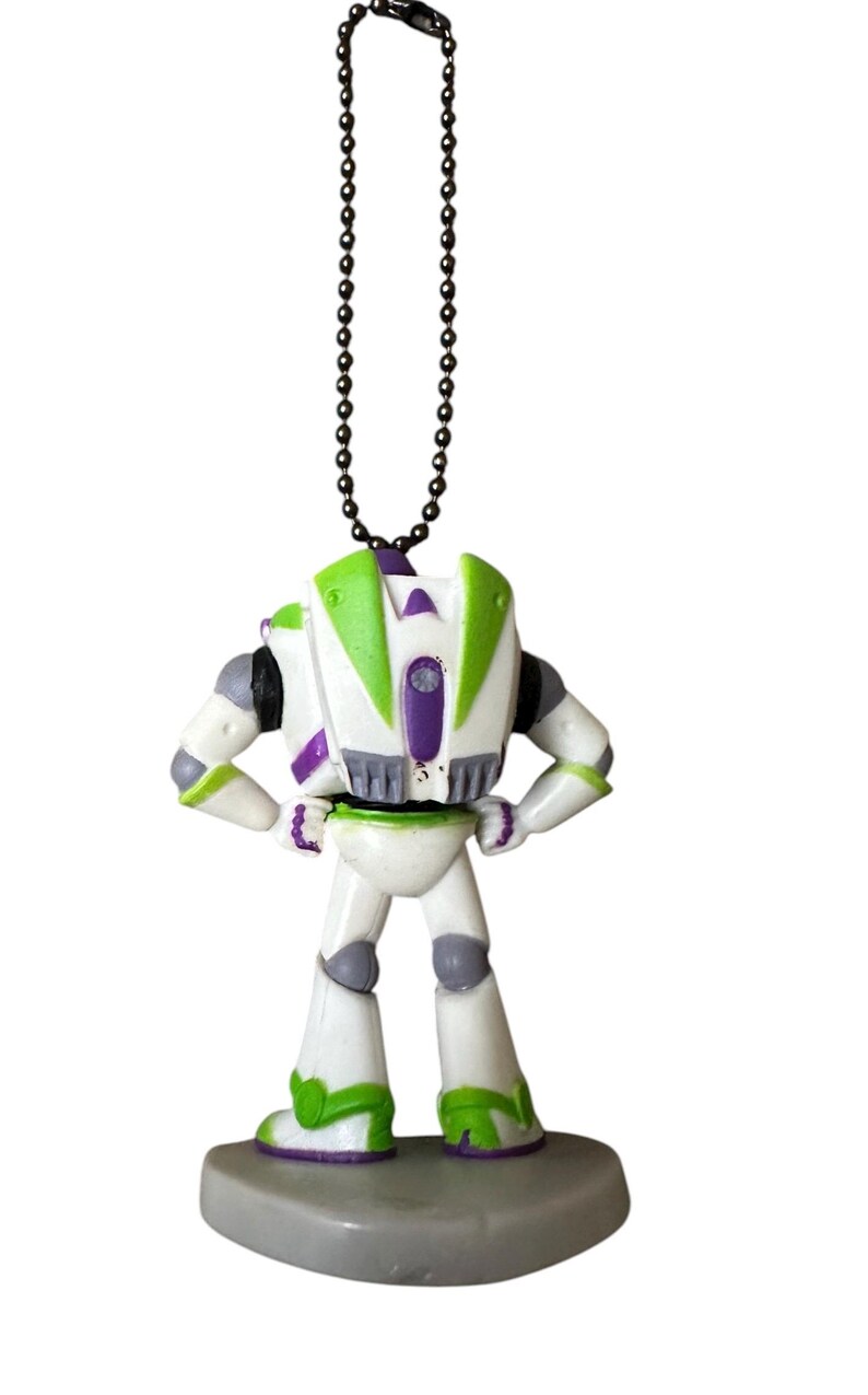Puede incluir: Una figura de Buzz Lightyear, vista desde atr&aacute;s, de pie sobre una base gris. El juguete presenta un cuerpo blanco con detalles verdes y morados, y una cadena negra para colgar. La figura tiene los brazos en las caderas.