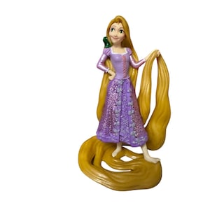 May include: A figurine of a princess with long blonde hair, wearing a purple dress with glitter accents. A small green chameleon sits on her shoulder. The figurine is standing on a base of her flowing hair.