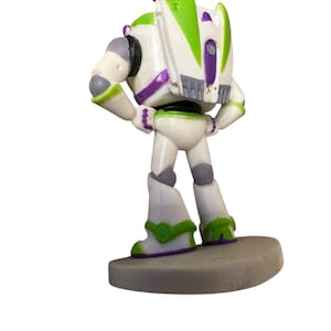 May include: A Buzz Lightyear pendant necklace. The pendant is a detailed figurine of the character, featuring white, green, and purple accents. The necklace has a brown cord. The pendant is standing on a gray base.