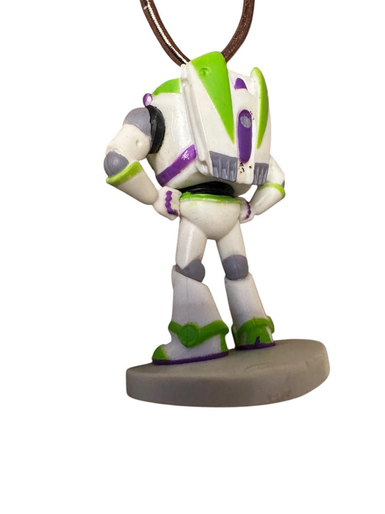 May include: A Buzz Lightyear pendant necklace. The pendant is a detailed figurine of the character, featuring white, green, and purple accents. The necklace has a brown cord. The pendant is standing on a gray base.