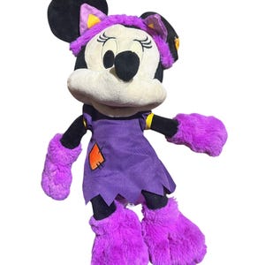 K&ouml;nnte beinhalten: Ein Pl&uuml;sch Minnie Mouse-Puppe in einem Halloween-Kost&uuml;m. Die Puppe hat ein lila Kleid mit einem orangefarbenen Aufn&auml;her, passende flauschige lila Handschuhe und Stiefel sowie einen lila Hexenhut mit gelben Akzenten. Minnie's schwarze Ohren und Schleife sind sichtbar.