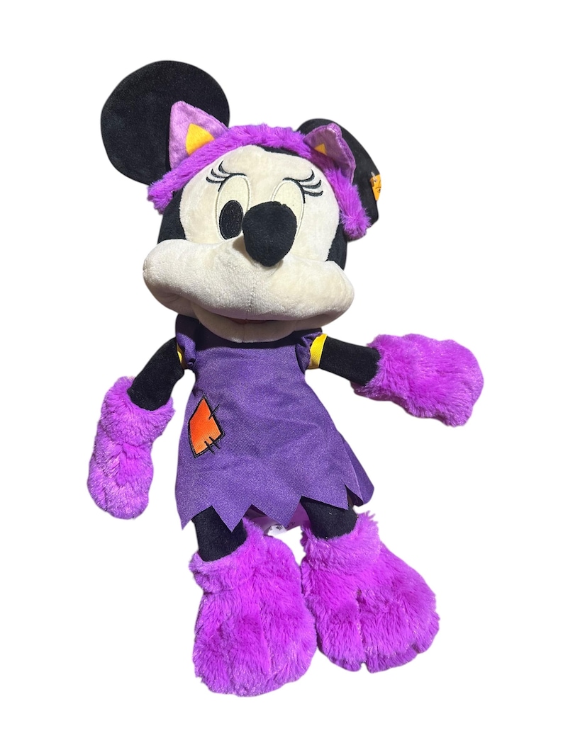K&ouml;nnte beinhalten: Ein Pl&uuml;sch Minnie Mouse-Puppe in einem Halloween-Kost&uuml;m. Die Puppe hat ein lila Kleid mit einem orangefarbenen Aufn&auml;her, passende flauschige lila Handschuhe und Stiefel sowie einen lila Hexenhut mit gelben Akzenten. Minnie's schwarze Ohren und Schleife sind sichtbar.