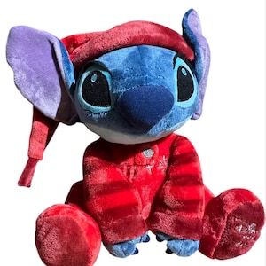 May include: A plush toy of the character Stitch, featuring blue fur, large purple-lined ears, and a dark blue nose. The character wears a red nightcap and matching red pajamas with horizontal stripes, a silver button, and a small embroidered star.