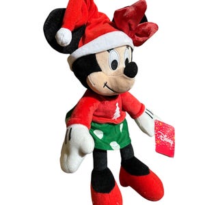 May include: A Minnie Mouse plush toy dressed for Christmas. She wears a red Santa hat, a red sweater with a Christmas tree design, and a green skirt with white polka dots. Her shoes and bow are red, and she has white gloves.
