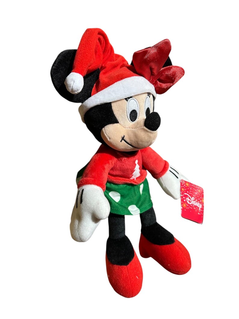 May include: A Minnie Mouse plush toy dressed for Christmas. She wears a red Santa hat, a red sweater with a Christmas tree design, and a green skirt with white polka dots. Her shoes and bow are red, and she has white gloves.