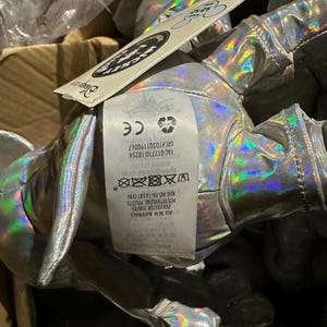 Puede incluir: Un peluche plateado hologr&aacute;fico con una etiqueta adjunta. La etiqueta incluye el texto "Mickey" y "Disney". El peluche est&aacute; hecho de fibras de poli&eacute;ster y gr&aacute;nulos de polietileno.