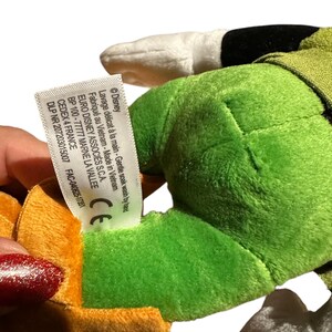 Pu&ograve; includere: Un peluche con colori verde, arancione e bianco. Il giocattolo ha un'etichetta con testo tra cui "Disney" e "Made in Vietnam". Il giocattolo &egrave; tenuto da una persona.