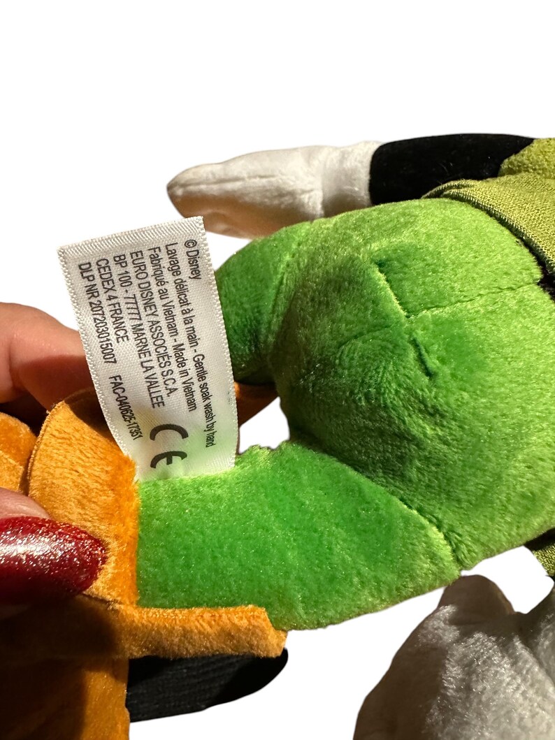 Pu&ograve; includere: Un peluche con colori verde, arancione e bianco. Il giocattolo ha un'etichetta con testo tra cui "Disney" e "Made in Vietnam". Il giocattolo &egrave; tenuto da una persona.
