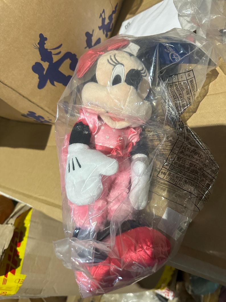 Puede incluir: Una mu&ntilde;eca de peluche de Minnie Mouse en una bolsa de pl&aacute;stico transparente. La mu&ntilde;eca lleva un vestido rosa, zapatos rojos y un lazo rojo. La mu&ntilde;eca tiene orejas negras y una cara blanca con nariz y ojos negros. Las manos de la mu&ntilde;eca son blancas con guantes negros.