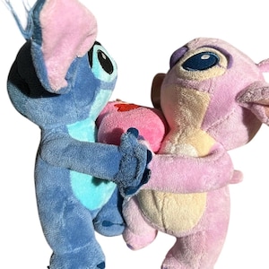 May include: Two plush toys, one blue and one pink, facing each other and holding a small pink heart between them. The blue plush has large ears with pink inner sections and dark eyes, while the pink plush features lighter pink accents.