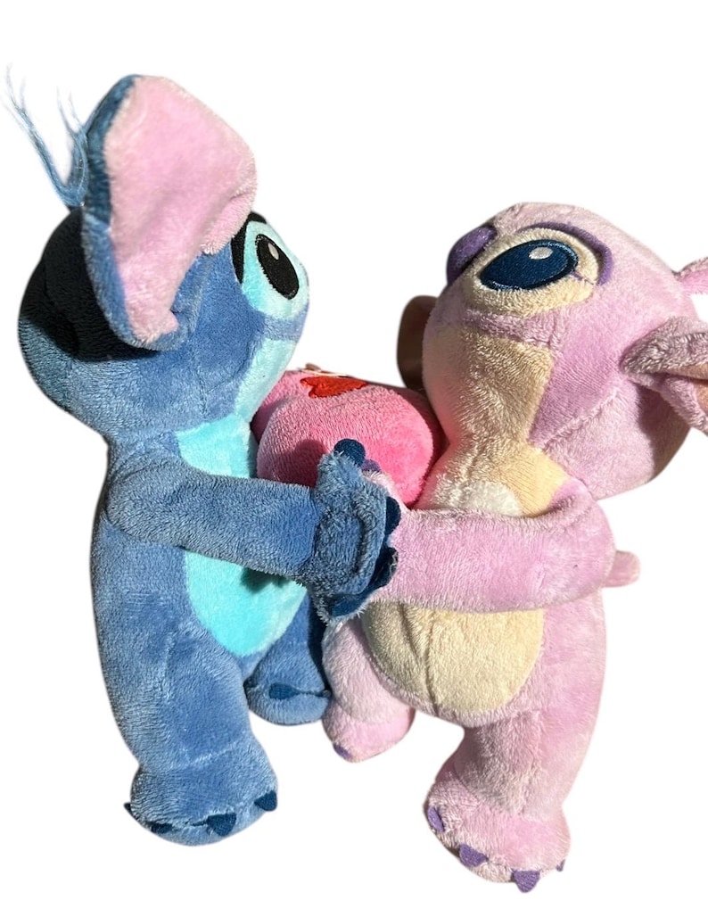 May include: Two plush toys, one blue and one pink, facing each other and holding a small pink heart between them. The blue plush has large ears with pink inner sections and dark eyes, while the pink plush features lighter pink accents.