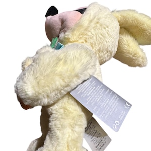 May include: A plush toy in the shape of a rabbit, with soft, cream-colored fur. The rabbit has pink inner ears, a black nose, and a small blue bow. The toy has a tag attached with text.
