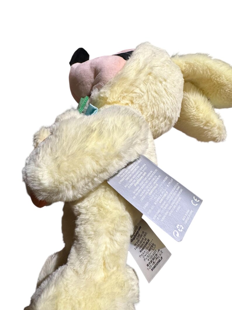 May include: A plush toy in the shape of a rabbit, with soft, cream-colored fur. The rabbit has pink inner ears, a black nose, and a small blue bow. The toy has a tag attached with text.