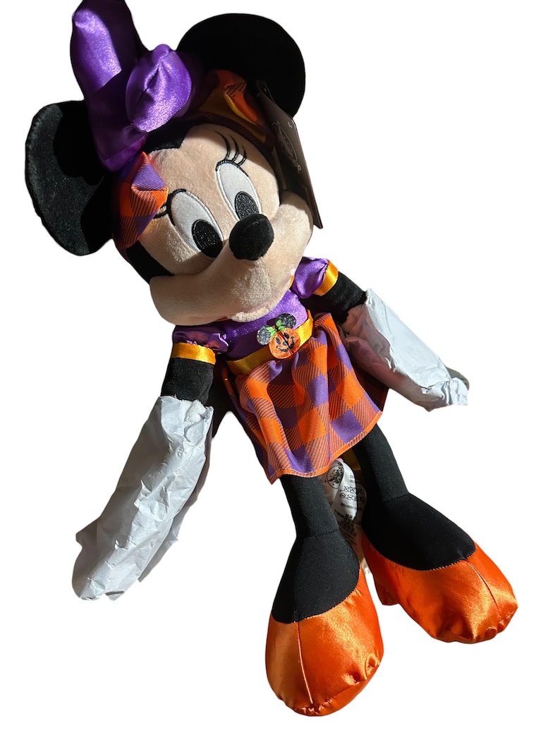 Op de afbeelding: Een Minnie Mouse pluche pop in een Halloween-kostuum. De pop heeft een zwarte kop en oren, een paarse strik en een geruite oranje en paarse jurk. De schoenen van de pop zijn oranje en ze heeft witte handschoenen.