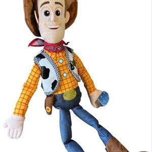 Op de afbeelding: Een pluche pop van een cowboyfiguur met een bruine hoed, een rode bandana, een geel en oranje geruit shirt met lange mouwen, een wit vest met zwarte koeienvlekken, een blauwe spijkerbroek en bruine laarzen met lichtgekleurde sporen.