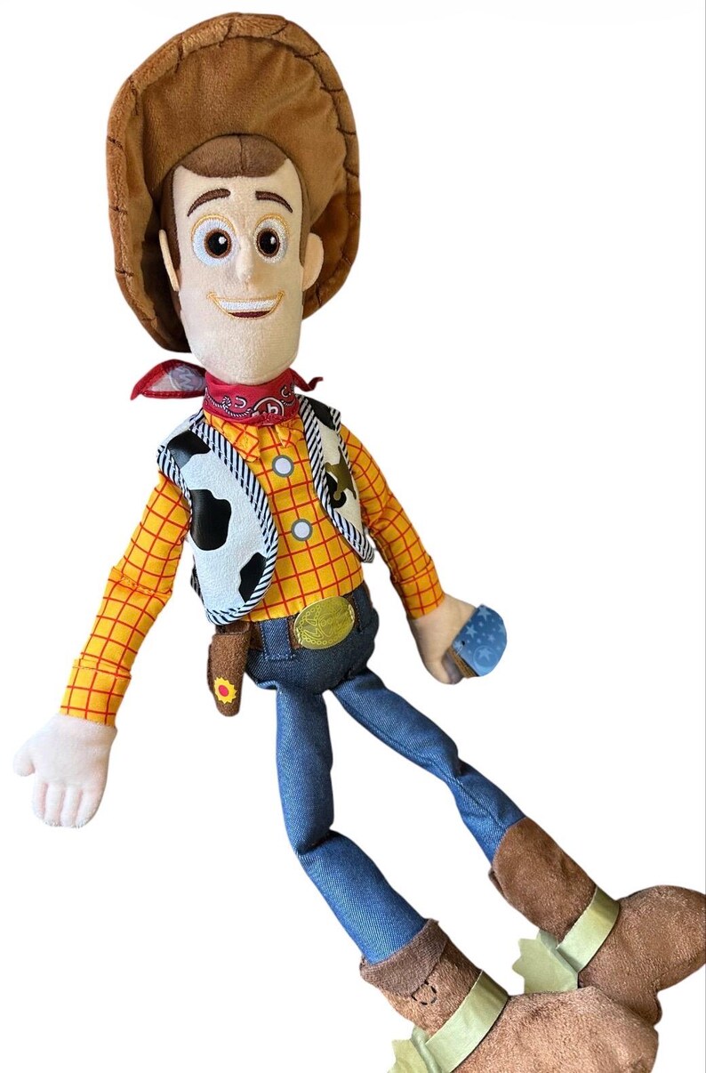 Op de afbeelding: Een pluche pop van een cowboyfiguur met een bruine hoed, een rode bandana, een geel en oranje geruit shirt met lange mouwen, een wit vest met zwarte koeienvlekken, een blauwe spijkerbroek en bruine laarzen met lichtgekleurde sporen.