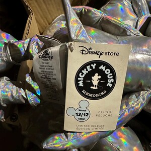 Puede incluir: Un mont&oacute;n de peluches plateados con reflejos iridiscentes. Se ve una etiqueta de Disney Store, con el logotipo de Mickey Mouse Memories y el texto "Serie 12/12". La etiqueta tambi&eacute;n indica "Edici&oacute;n Limitada".
