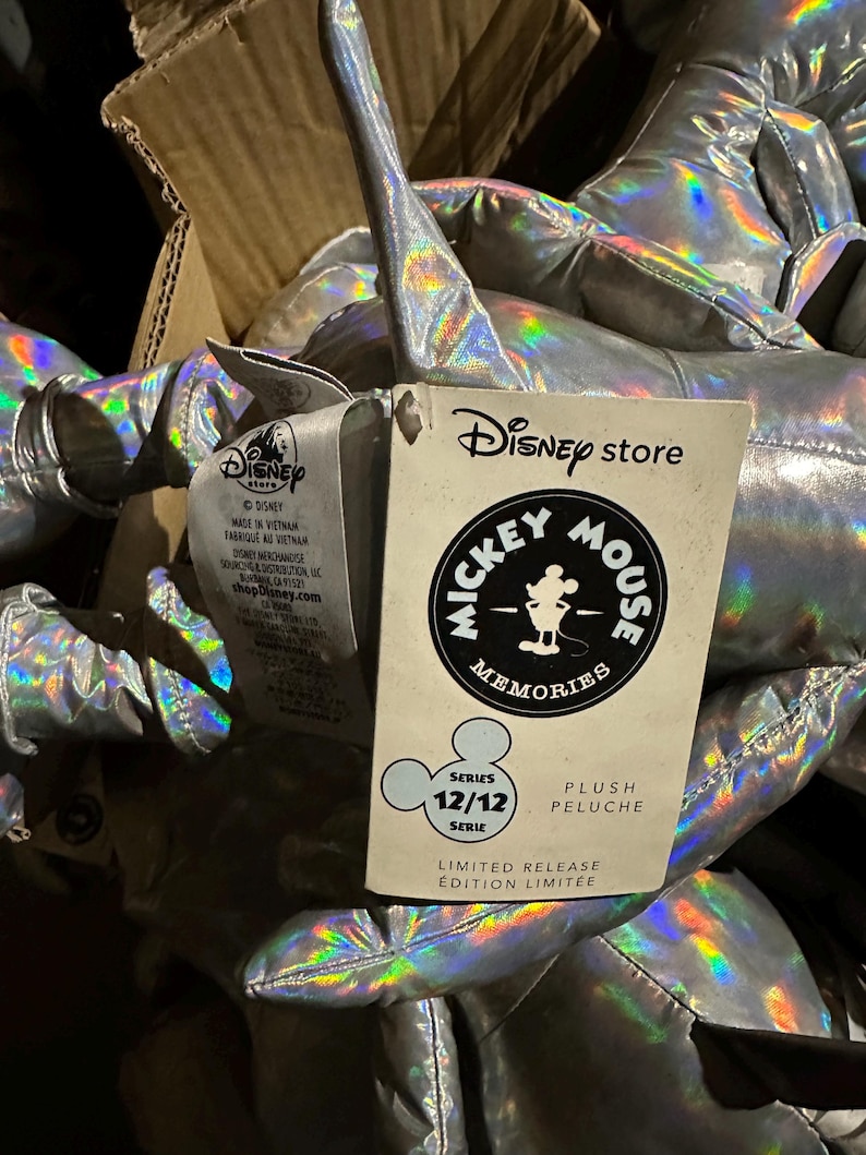 Puede incluir: Un mont&oacute;n de peluches plateados con reflejos iridiscentes. Se ve una etiqueta de Disney Store, con el logotipo de Mickey Mouse Memories y el texto "Serie 12/12". La etiqueta tambi&eacute;n indica "Edici&oacute;n Limitada".