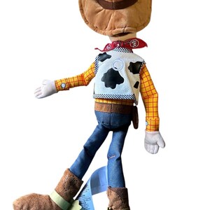Op de afbeelding: Een pluche pop van een cowboyfiguur met een bruine hoed, een rode bandana, een wit vest met zwarte koeienvlekken, een geel shirt met lange mouwen en een rood ruitpatroon, een blauwe broek en bruine laarzen. Een blauw productlabel zit aan de laars.