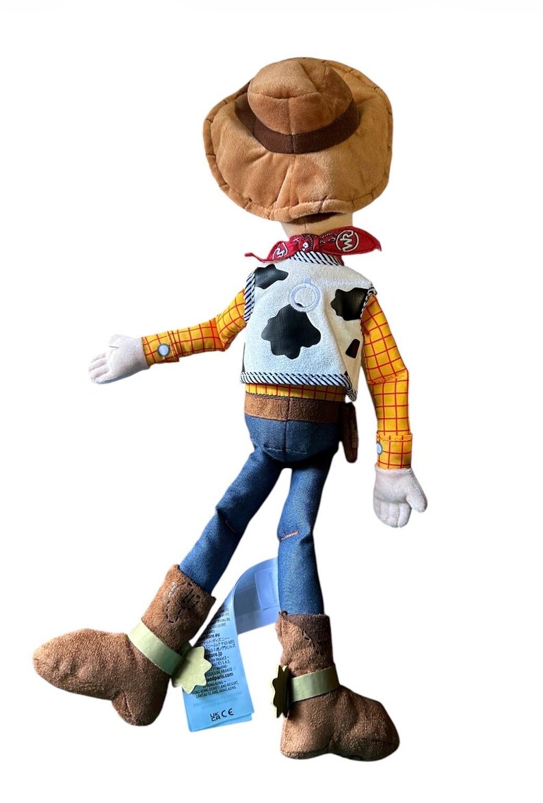 Op de afbeelding: Een pluche pop van een cowboyfiguur met een bruine hoed, een rode bandana, een wit vest met zwarte koeienvlekken, een geel shirt met lange mouwen en een rood ruitpatroon, een blauwe broek en bruine laarzen. Een blauw productlabel zit aan de laars.