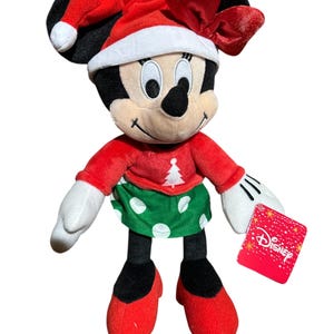 May include: A Minnie Mouse plush toy dressed for Christmas. She wears a red Santa hat with a white trim and a matching bow. Her red top has a Christmas tree design, and she has a green skirt with white polka dots. The toy has a Disney tag.