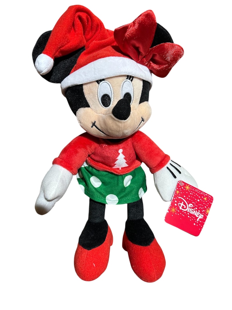 May include: A Minnie Mouse plush toy dressed for Christmas. She wears a red Santa hat with a white trim and a matching bow. Her red top has a Christmas tree design, and she has a green skirt with white polka dots. The toy has a Disney tag.